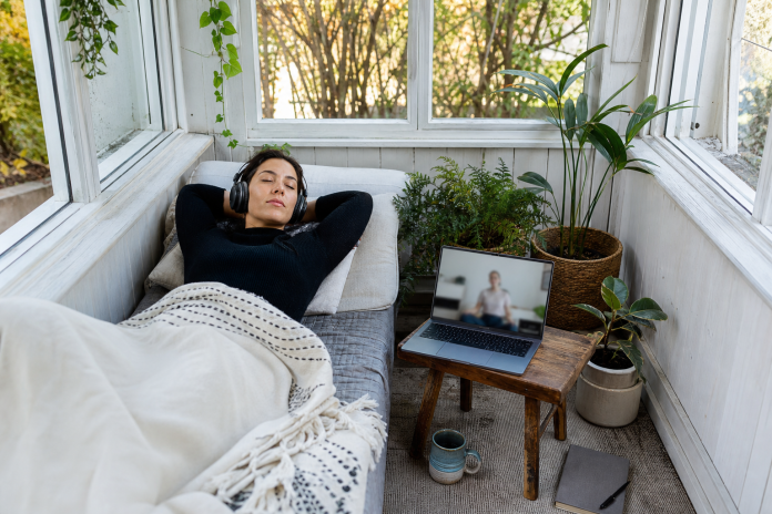 Online session from a sunroom daybed Woman lying on a daybed in a sunroom with a laptop open on a stool beside her showing a video call, headphones on