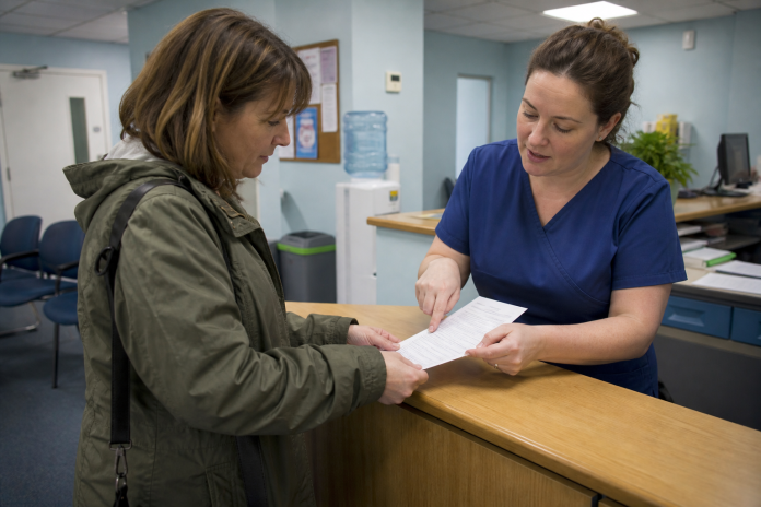 Asking questions at a clinic reception Woman reading a printed leaflet at a clinic reception desk while a receptionist points to a page, waiting-room chairs behind them
