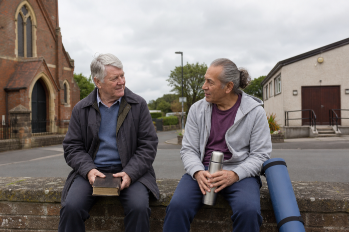 Conversation between a church and a community hall Two older men talking on a bench outside a small brick church and a community hall, one with a bible in his lap, the other with a folded yoga mat