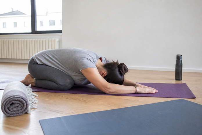 Child's pose in a quiet studio Woman in a bright studio kneeling in child's pose on a yoga mat with her hands folded under her forehead, another mat and a water bottle nearby