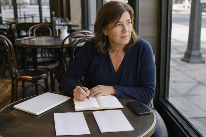 Woman at a cafe table comparing two brochures side by side with a pen and a notebook, coffee cup and phone nearby
