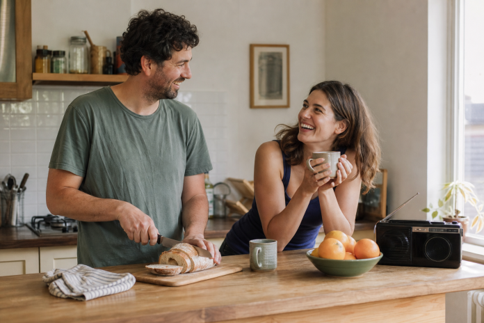 Easy company at the kitchen counter Two friends laughing over coffee at a kitchen counter, one leaning on the counter with a mug and the other slicing bread, morning light