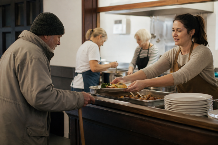 Serving at a community kitchen counter Volunteer in an apron handing a plate across a community kitchen counter to an older man, other helpers working in the background