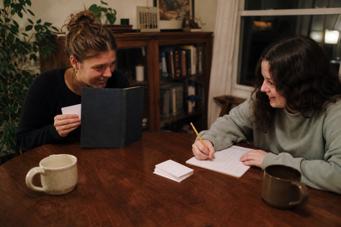 Two friends at a dining table with a small deck of plain white cards, one holding a card face down behind a propped-up book as a screen, the other writing on a scoresheet