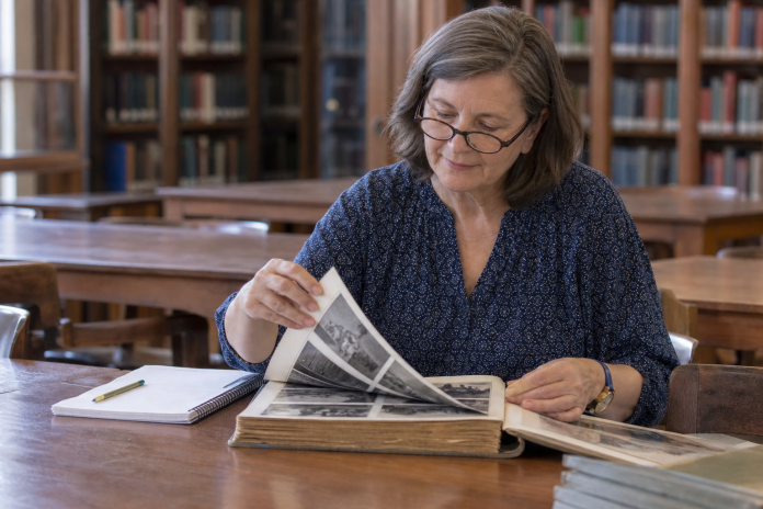 Turning the pages of an old photograph album Woman in a library reading room turning the pages of an old bound volume of photographs, reading glasses on, soft window light