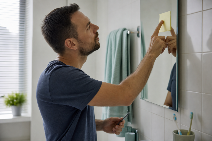 A note on the bathroom mirror Man writing a short note on a sticky pad and pressing it to the edge of a bathroom mirror, morning light, toothbrush and towel nearby
