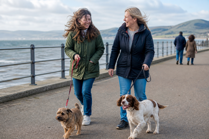 Laughing on a breezy seafront walk Woman laughing with a friend while walking two dogs along a seafront promenade on a bright breezy day