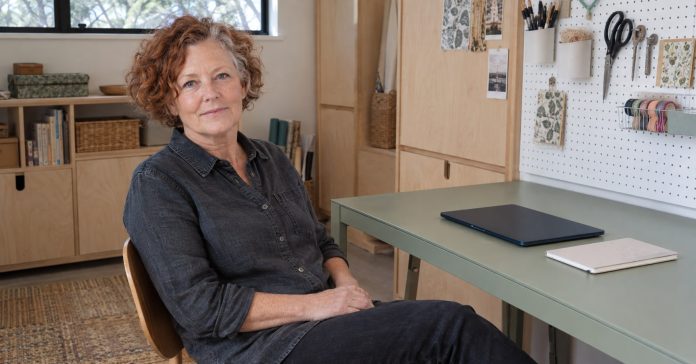 Woman in her sixties pausing at a green metal desk with a closed modern navy laptop in a home craft room.