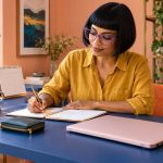 Can Reiki Attract Money and Love? What Intention-Setting Actually Does A woman writes a weekly plan beside a closed pink laptop.