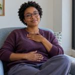 The Benefits of Reiki Healing: A Short Summary and Where to Read More A woman sits comfortably in a blue armchair, resting one hand on her chest and one on her abdomen.