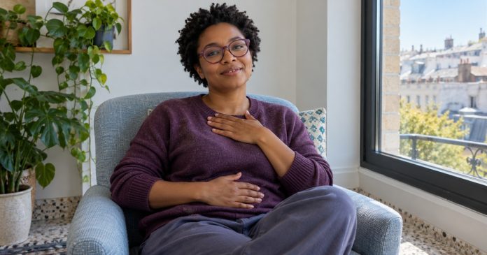 The Benefits of Reiki Healing: A Short Summary and Where to Read More — editorial image A woman sits comfortably in a blue armchair, resting one hand on her chest and one on her abdomen.