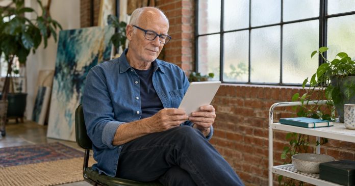 An older man in glasses reads an unbranded e-reader in a bright brick-walled studio.