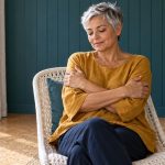 Reiki, the Japanese “Energy Healing Therapy”: A Short Definition and Where to Read More A silver-haired woman sits fully clothed in a white woven chair, resting her hands on her upper arms.