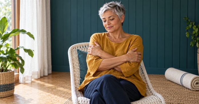 Reiki, the Japanese "Energy Healing Therapy": A Short Definition and Where to Read More — editorial image A silver-haired woman sits fully clothed in a white woven chair, resting her hands on her upper arms.