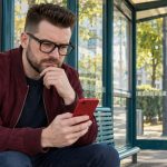 Toxic Relationships: The Signs, the Line Between Toxic and Abusive, and How to Step Back A man sits at an open-air stop holding a red smartphone.