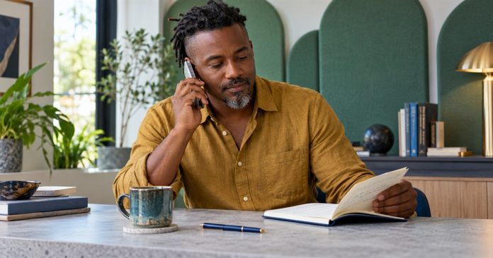 A man checks a question notebook while speaking on a phone.