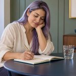 Three Aura-Cleansing Practices: Breathing, Writing and Scent A woman writes in a notebook beside a glass of water.