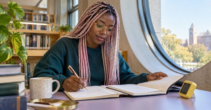 A woman compares notes in an open journal at a library desk.