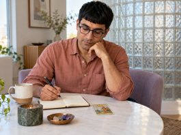 The Psychology of Tarot: Mental Wellness and Reflection in 2026 A man journals beside one tarot card at a white pedestal table.