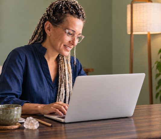 What Does Research Say About Reiki? Evidence and Limits A Reiki practitioner with glasses and highlighted braids looking at her laptop in a calm room with sage walls.