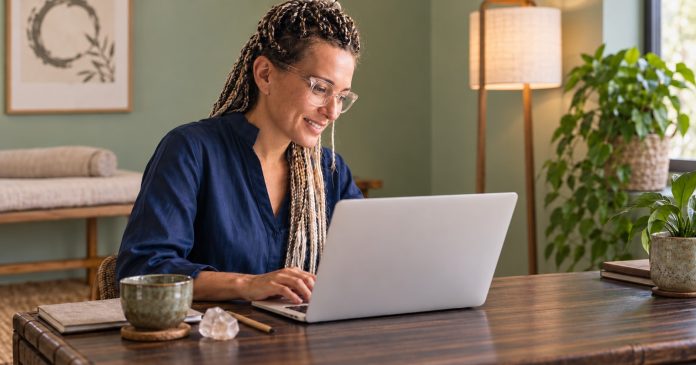 Reiki Research — Reading on a Laptop A Reiki practitioner with glasses and highlighted braids looking at her laptop in a calm room with sage walls.